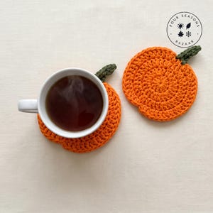 Op de afbeelding: Twee oranje gehaakte onderzetters in de vorm van pompoenen met groene steeltjes. Eén onderzetter ondersteunt een witte keramische mok gevuld met donkere vloeistof. Een rond logo in de rechterbovenhoek toont Four Seasons Bazaar met iconen.