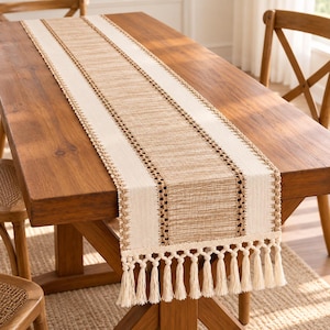 May include: A rectangular table runner featuring a central tan woven panel flanked by cream-colored borders and intricate brown macrame-style detailing. The ends are finished with a row of long, cream-colored knotted fringe tassels.