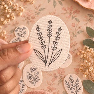 May include: An oval-shaped sticker featuring a black line drawing of three lavender stalks with leaves on a textured off-white background. The sticker is held up, displaying the botanical illustration clearly against the paper surface.