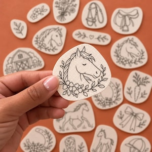May include: A collection of off-white, irregular-shaped stickers featuring black line art illustrations of horses, barns, saddles, bridles, flowers, carrots, and cowboy boots. Each sticker has a textured surface and a simple, clean design.