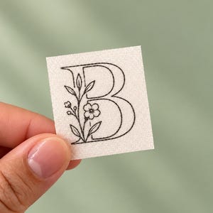 May include: A square white patch featuring a black embroidered letter B. The letter is decorated with a delicate black line art illustration of a small flower and leaves growing along the left side of the character, creating a botanical design.