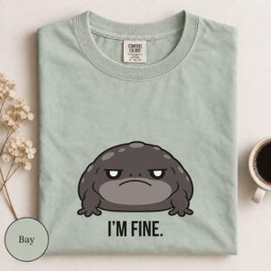 Puede incluir: Una camiseta de manga corta color verde salvia con un gráfico de un sapo gris oscuro de aspecto gruñón y el texto I'M FINE. impreso en negro debajo. La prenda tiene cuello redondo y una etiqueta de la marca Comfort Colors en el interior.