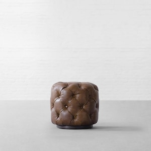 May include: A round, brown leather ottoman featuring a tufted design with deep button accents across the top and sides. The piece is supported by a dark, circular base, providing a stable foundation for the upholstered, textured leather surface.