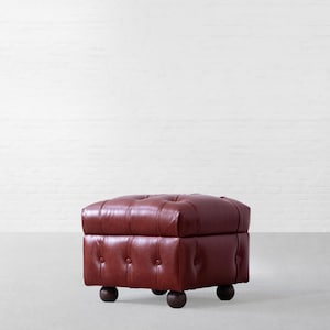 May include: A square, deep red leather ottoman featuring a tufted design with fabric-covered buttons on the top and sides. The piece is supported by four dark, rounded wooden feet, providing a low-profile structure with a classic, boxy shape.