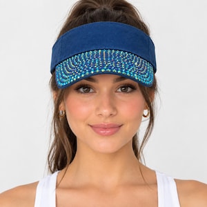 Pode incluir: Uma viseira azul-marinho com aba curva densamente adornada com strass iridescentes multicoloridos dispostos num padrão radiante. A banda da coroa é feita de tecido azul-marinho sólido com um design estruturado e ajustável.