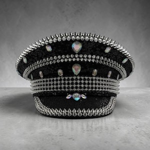 Könnte beinhalten: Eine schwarze Mütze im Militärstil, bedeckt mit schimmernden Pailletten, verziert mit Reihen silberfarbener Metallspikes und irisierenden, tropfenförmigen Kristallen, die über die Krone, das Band und den geschwungenen Schirm verteilt sind.