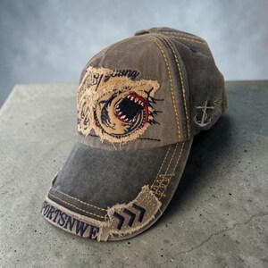 May include: A distressed brown baseball cap featuring a frayed patch with an embroidered shark graphic and the text "Young". The brim displays the text "SPORTSNWE" in dark blue lettering, and the side of the cap includes an embroidered anchor icon.