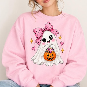 Op de afbeelding: Een lichtroze sweater met ronde hals en een centrale afbeelding van een witte geest met grote zwarte ogen, roze wangen en een grote roze strik met stippen op het hoofd. De geest houdt een oranje pompoenemmer vast gevuld met kleurrijke snoepjes.