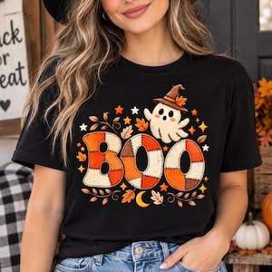 Op de afbeelding: Een zwart t-shirt met een grafische print van het woord BOO in een patchwork oranje en crèmekleurig patroon. Boven de tekst staat een klein wit spookje met een bruine heksenhoed. Het ontwerp is omringd door oranje sterren, herfstbladeren en een maan.