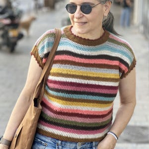 Könnte beinhalten: Ein bunt gestreiftes Häkeltop mit kurzen Ärmeln. Das Oberteil hat horizontale Bänder in verschiedenen Farben, darunter Braun, Blau, Rosa, Gelb und Grün. Der Halsausschnitt und die Ärmelränder sind braun eingefasst. Das Oberteil wird mit einer blauen Jeans getragen.