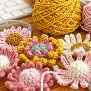 May include: A collection of crocheted flowers in various colors, including pink, white, yellow, and gray. The flowers are arranged on a wooden surface.