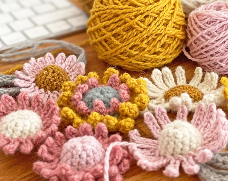 May include: A collection of crocheted flowers in various colors, including pink, white, yellow, and gray. The flowers are arranged on a wooden surface.