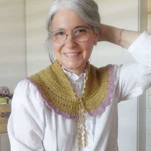 Op de afbeelding: Een gehaakte kraag met een gegolfde rand in mosterdgeel en mauve. De kraag heeft een strik aan de voorkant.