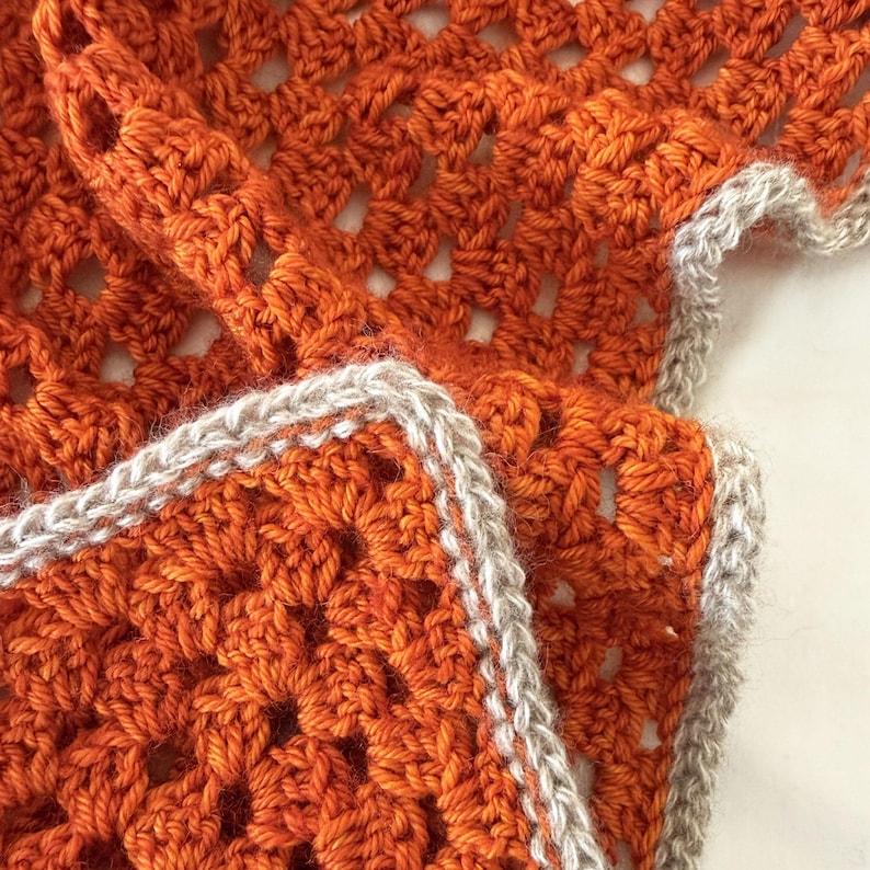 Puede incluir: Primer plano de una manta de ganchillo hecha a mano. La manta presenta un color naranja vibrante con un patr&oacute;n calado detallado. Los bordes est&aacute;n acabados con un borde gris claro contrastante, agregando un toque de elegancia al textil acogedor.