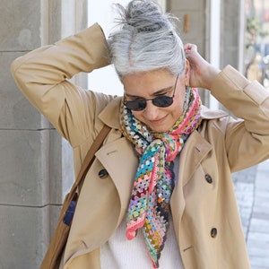 May include: A woman wearing a beige trench coat with a colorful crocheted scarf. The scarf is a mix of pink, blue, green, and brown yarn.