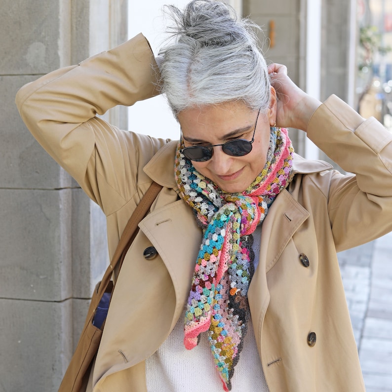 May include: A woman wearing a beige trench coat with a colorful crocheted scarf. The scarf is a mix of pink, blue, green, and brown yarn.