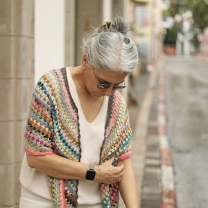 May include: A woman wearing a colorful crocheted shawl with a white top and gray hair. She is wearing a watch on her left wrist.