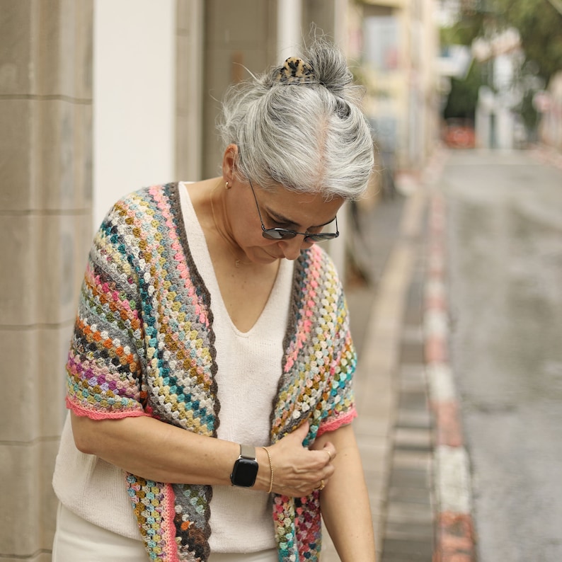 May include: A woman wearing a colorful crocheted shawl with a white top and gray hair. She is wearing a watch on her left wrist.