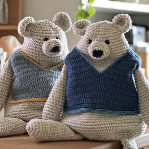 Puede incluir: Dos osos de peluche tejidos a crochet que llevan chalecos azules y grises con ribetes amarillos. Los osos están sentados uno al lado del otro sobre una superficie de madera.