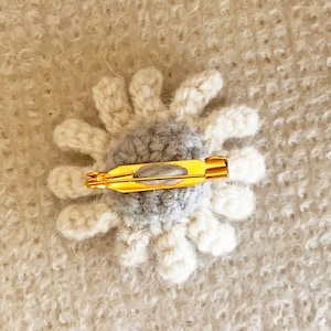 May include: A crocheted flower brooch with a gray center and white petals. The brooch has a gold metal pin on the back.