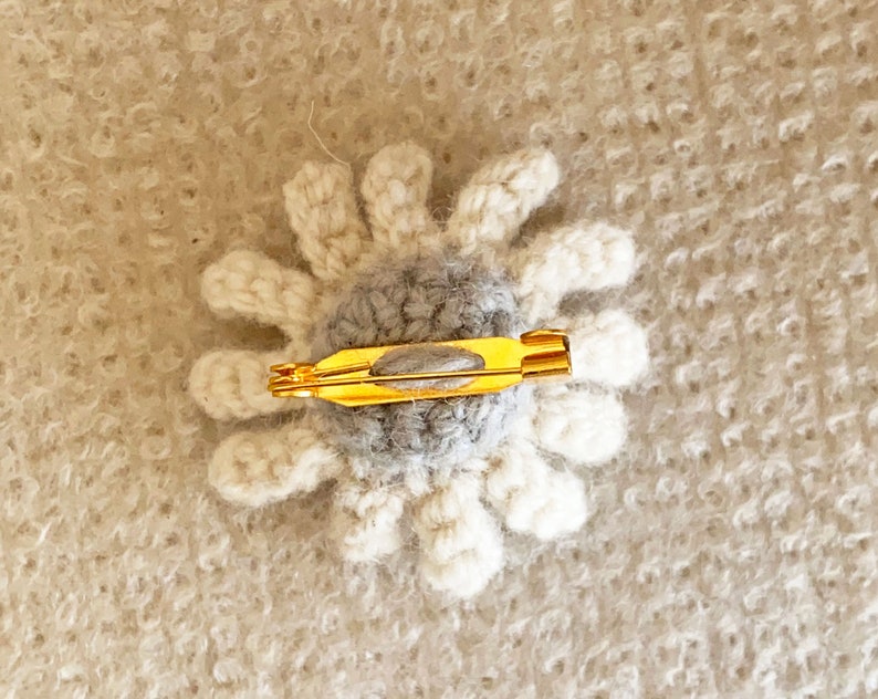 May include: A crocheted flower brooch with a gray center and white petals. The brooch has a gold metal pin on the back.