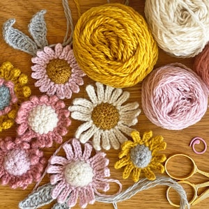 May include: A collection of crocheted flowers in various colors, including pink, yellow, and white, with gray leaves. The flowers are arranged on a wooden surface with balls of yarn in pink, white, and yellow. There are also gold scissors and a pink crochet hook.