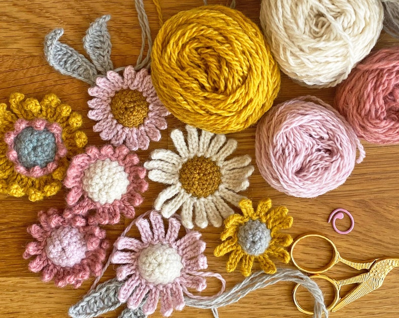 May include: A collection of crocheted flowers in various colors, including pink, yellow, and white, with gray leaves. The flowers are arranged on a wooden surface with balls of yarn in pink, white, and yellow. There are also gold scissors and a pink crochet hook.