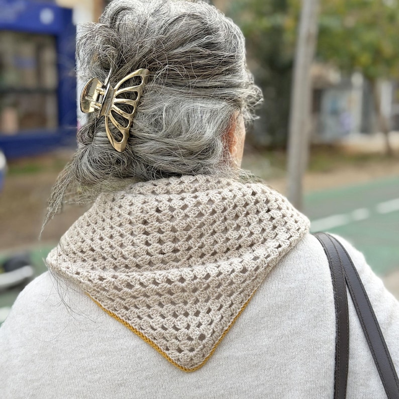 Puede incluir: Una mujer con cabello gris lleva una pinza dorada para el pelo con forma de mariposa y un chal beige de ganchillo con borde amarillo. El chal est&aacute; drapeado sobre los hombros.