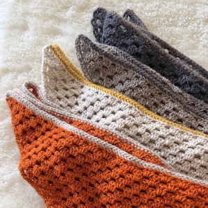 Puede incluir: Primer plano de varias bufandas de ganchillo en varios colores. Las bufandas est&aacute;n dispuestas en diagonal, mostrando tonos de naranja, crema, gris y gris oscuro. Cada bufanda tiene un patr&oacute;n texturizado y un borde contrastante.