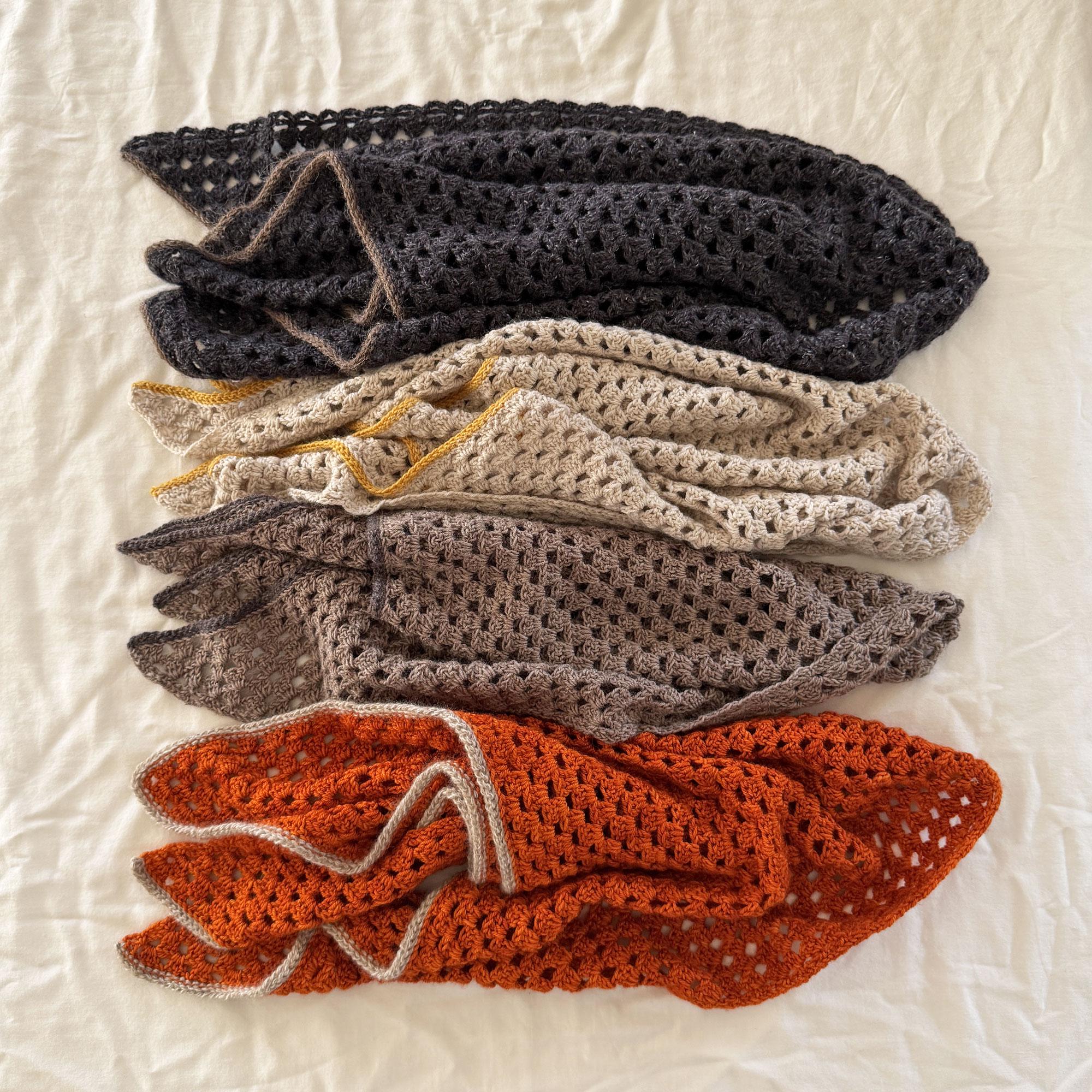 Puede incluir: Cuatro mantas de ganchillo apiladas. Las mantas son en tonos de gris, beige, marr&oacute;n y naranja. Cada manta tiene un ribete de diferente color. Las mantas est&aacute;n dobladas y dispuestas sobre una superficie blanca.