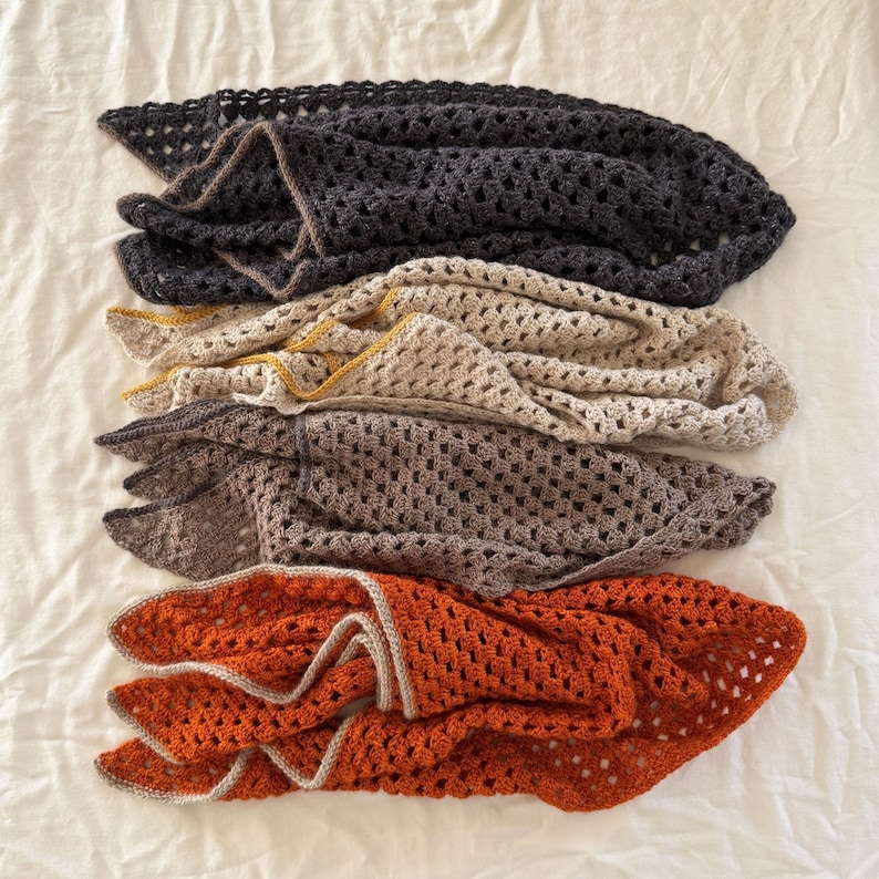Puede incluir: Cuatro mantas de ganchillo apiladas. Las mantas son en tonos de gris, beige, marr&oacute;n y naranja. Cada manta tiene un ribete de diferente color. Las mantas est&aacute;n dobladas y dispuestas sobre una superficie blanca.