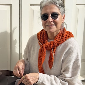 Puede incluir: Una bufanda naranja tejida a ganchillo con un ribete gris claro se lleva alrededor del cuello. La bufanda est&aacute; anudada por delante. La persona lleva un jersey gris claro y gafas de sol redondas negras.