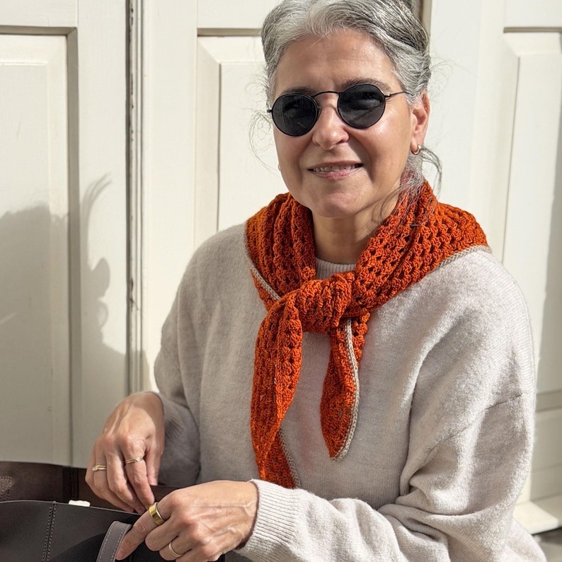 Puede incluir: Una bufanda naranja tejida a ganchillo con un ribete gris claro se lleva alrededor del cuello. La bufanda est&aacute; anudada por delante. La persona lleva un jersey gris claro y gafas de sol redondas negras.