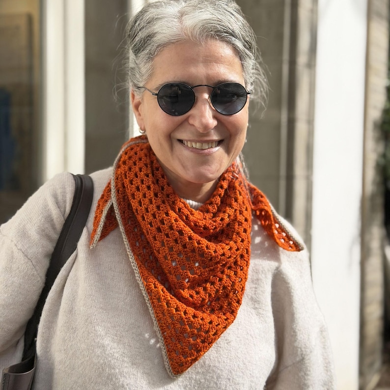 Puede incluir: Un chal de ganchillo naranja con forma triangular y borde gris claro. El chal se lleva alrededor del cuello y los hombros. El dise&ntilde;o de tejido abierto es visible. La persona de la imagen lleva gafas de sol y un su&eacute;ter de color claro.