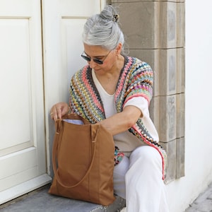 May include: A woman wearing a colorful crocheted shawl and white pants is holding a brown leather tote bag.