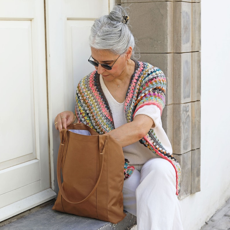 May include: A woman wearing a colorful crocheted shawl and white pants is holding a brown leather tote bag.