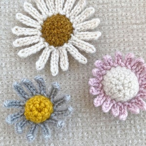 May include: Three crocheted flower embellishments in different colors. The first flower is white with a yellow center. The second flower is gray with a yellow center. The third flower is pink with a white center.