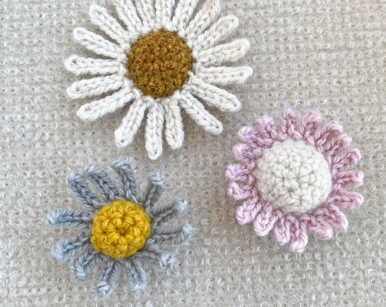 May include: Three crocheted flower embellishments in different colors. The first flower is white with a yellow center. The second flower is gray with a yellow center. The third flower is pink with a white center.