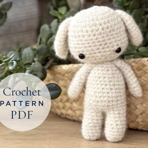 May include: A crocheted white amigurumi puppy with black eyes. The puppy is sitting on a woven basket with a white circle that says "Crochet Pattern PDF".