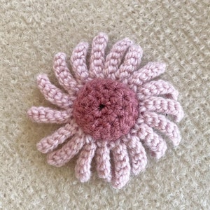 May include: A pink crocheted flower with a darker pink center. The flower is on a light brown fuzzy background.