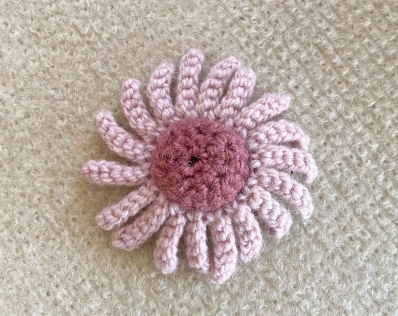 May include: A pink crocheted flower with a darker pink center. The flower is on a light brown fuzzy background.
