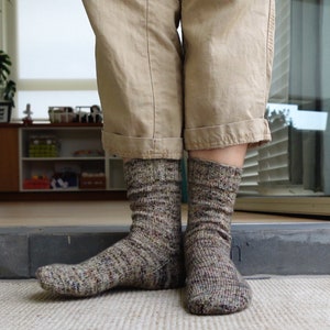 May include: A pair of hand-knitted socks in a speckled brown and gray yarn. The socks are worn with tan pants, showcasing the textured knit and color variations. The socks are designed for warmth and comfort.