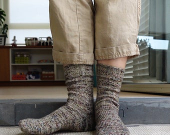 May include: A pair of hand-knitted socks in a speckled brown and gray yarn. The socks are worn with tan pants, showcasing the textured knit and color variations. The socks are designed for warmth and comfort.