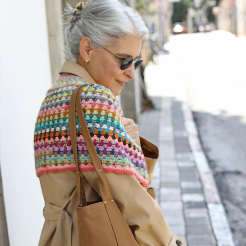 May include: A woman wearing a beige trench coat with a colorful crocheted shawl over her shoulders. She is carrying a brown leather tote bag.