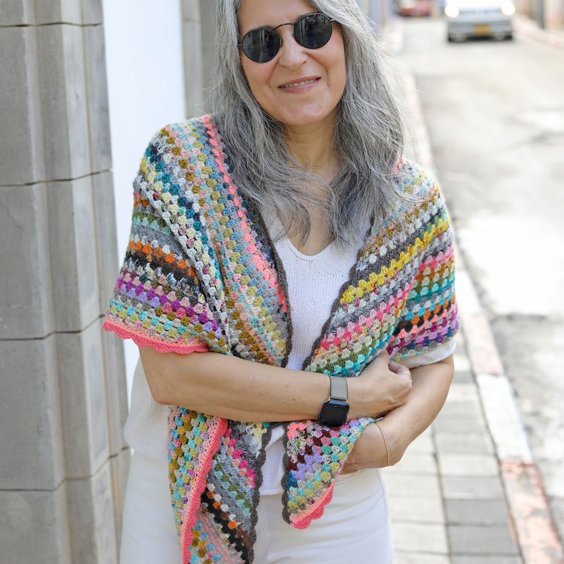 May include: A woman wearing a white shirt and white pants with a colorful crochet shawl. The shawl has a scalloped edge and is made with a variety of colors, including pink, blue, green, yellow, and purple.