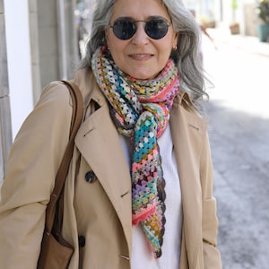 May include: A woman wearing a beige trench coat with a colorful crocheted scarf. The scarf is made of various colors, including pink, blue, green, and brown.