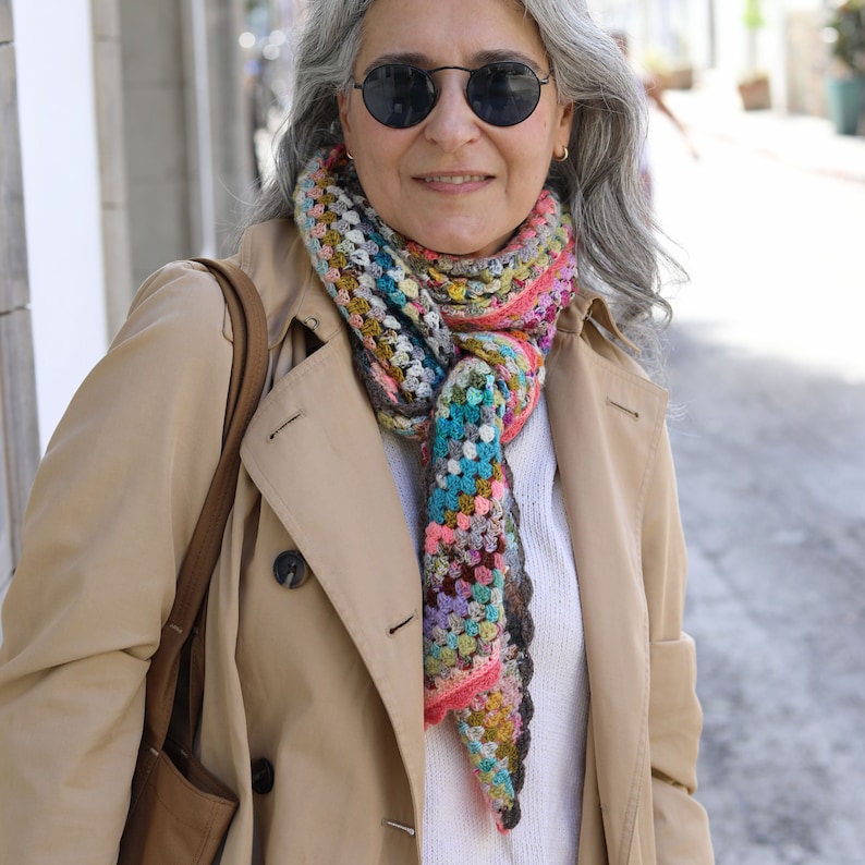 May include: A woman wearing a beige trench coat with a colorful crocheted scarf. The scarf is made of various colors, including pink, blue, green, and brown.