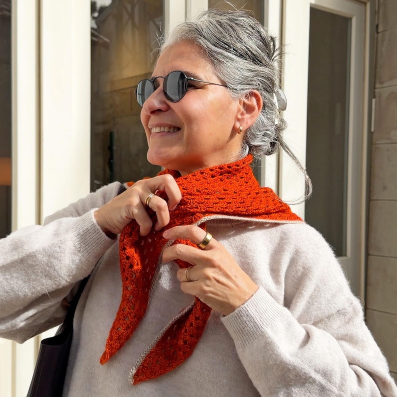 Puede incluir: Bufanda de ganchillo naranja con borde blanco, sobre un su&eacute;ter gris claro. La bufanda es triangular y tiene un dise&ntilde;o de tejido abierto y texturizado. La persona de la imagen lleva gafas de sol y anillos dorados.