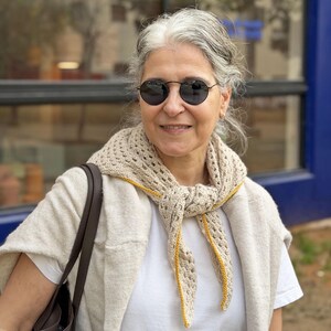 Puede incluir: Una mujer con una bufanda beige de ganchillo con ribete amarillo. La bufanda est&aacute; drapeada alrededor del cuello y los hombros. Tambi&eacute;n lleva una camiseta blanca, un su&eacute;ter beige y gafas de sol negras.