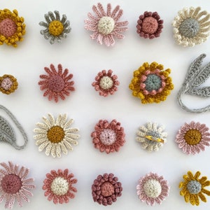 May include: A collection of 17 crocheted flower pins in various shades of pink, yellow, and gray. The flowers are all different sizes and shapes, and some have a center button.